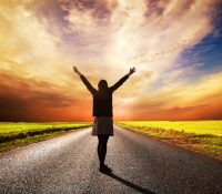 Happy woman standing with hands up on long straight road facing the sun. Sunset sky